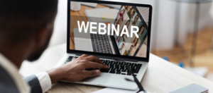 A person uses a laptop showing a video call, with the word “Webinar” on screen.